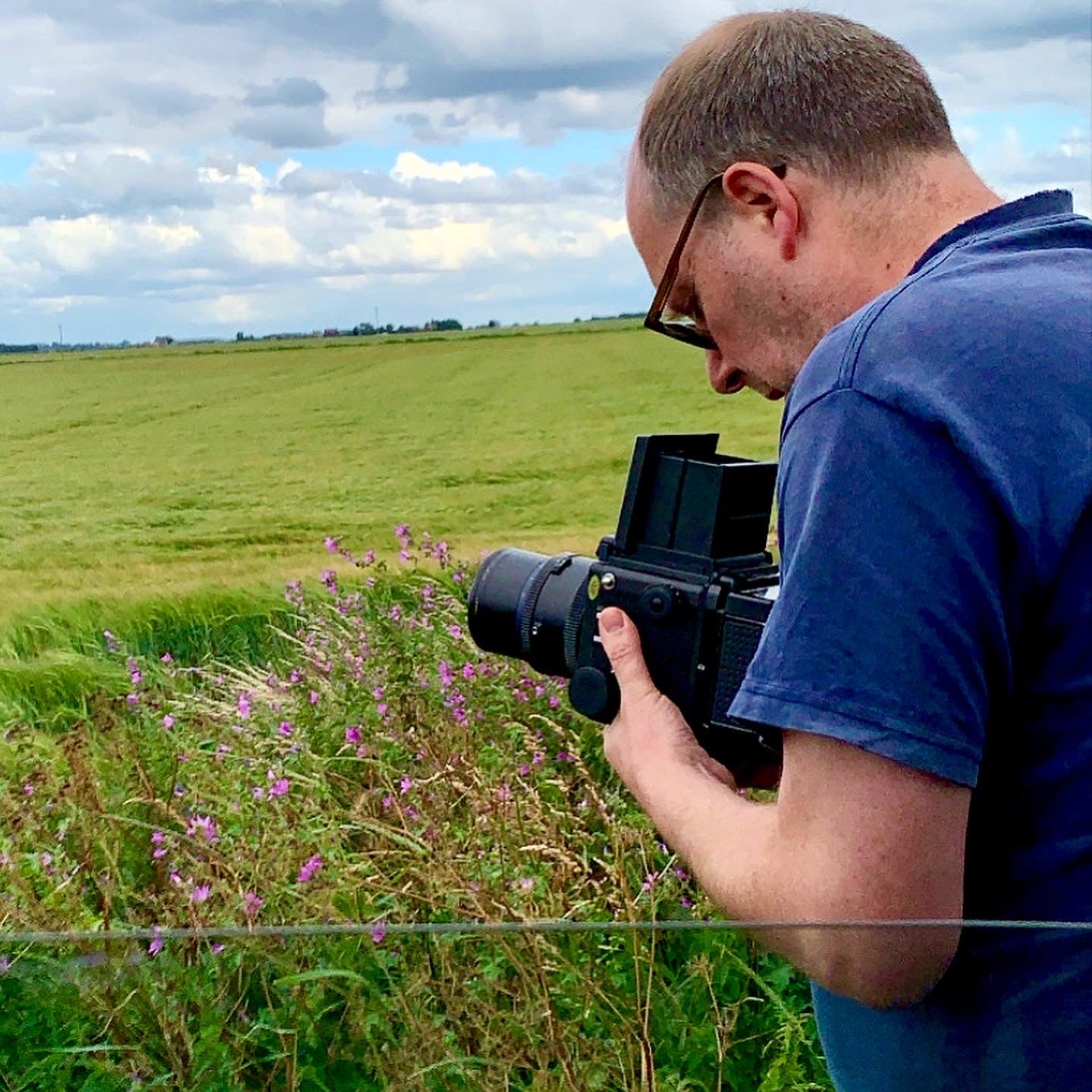 Richard Heeps with a Mamiya RB67 vintage camera in a green field with clouds in the sky. 