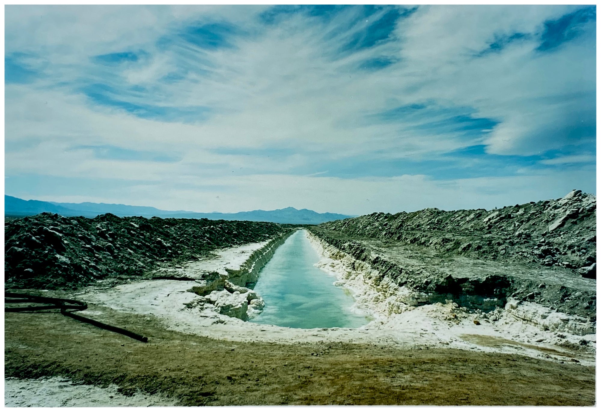 Photograph by Richard Heeps. A watery white trench is dug into the centre going into the distance with grey mounds of earth either side. The sky is vast and blue, with light clouds.