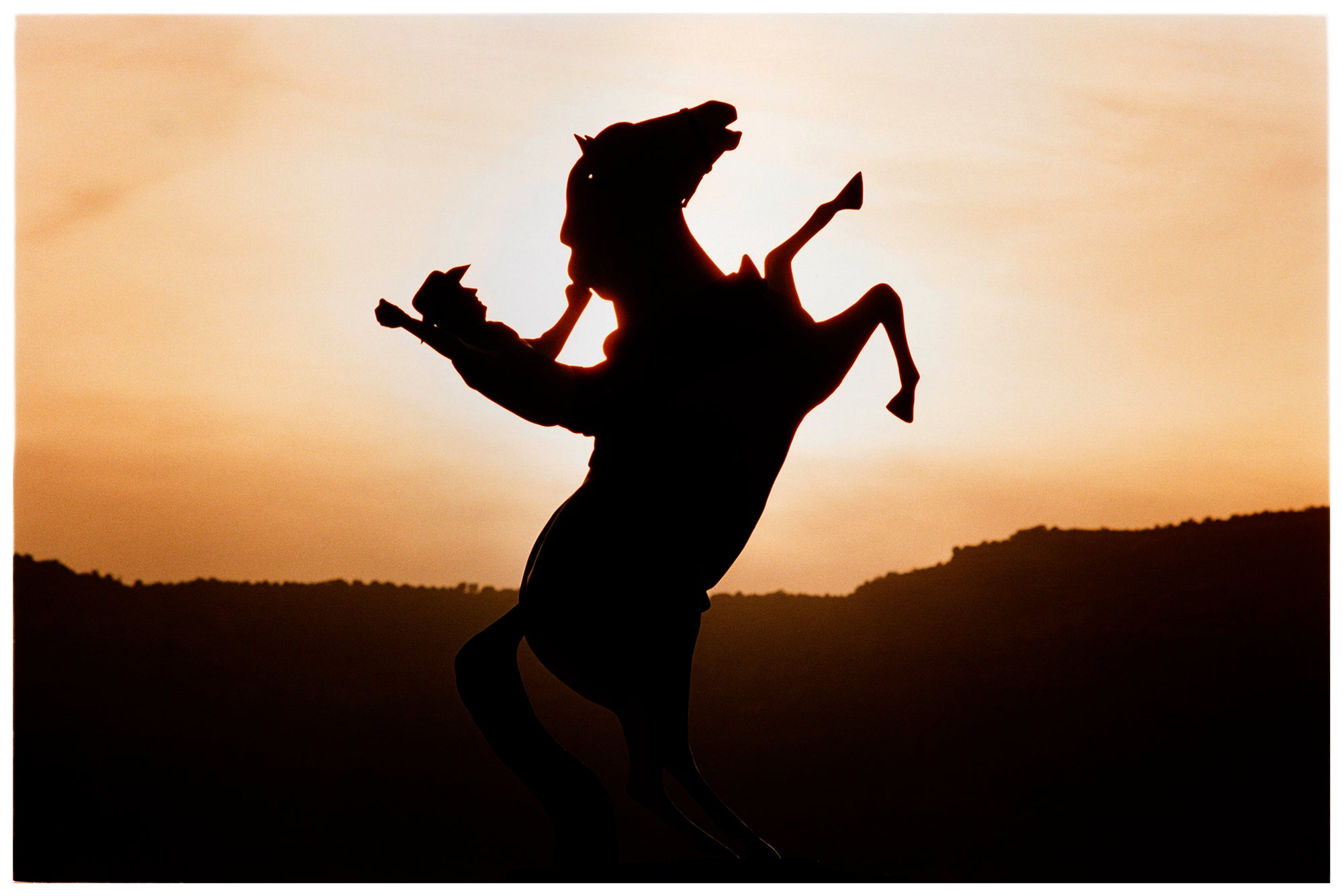 Photograph by Richard Heeps. The photograph shows a sunset and in the middle of the photograph is the silhouette of a statute of a cowboy on his bucking horse with one arm on the reins and the other held high.