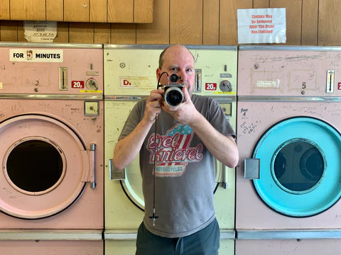 Richard Heeps with a Hasselblad Super wide vintage film camera in a London launderette with a backdrop of pink, yellow and blue washing machines.