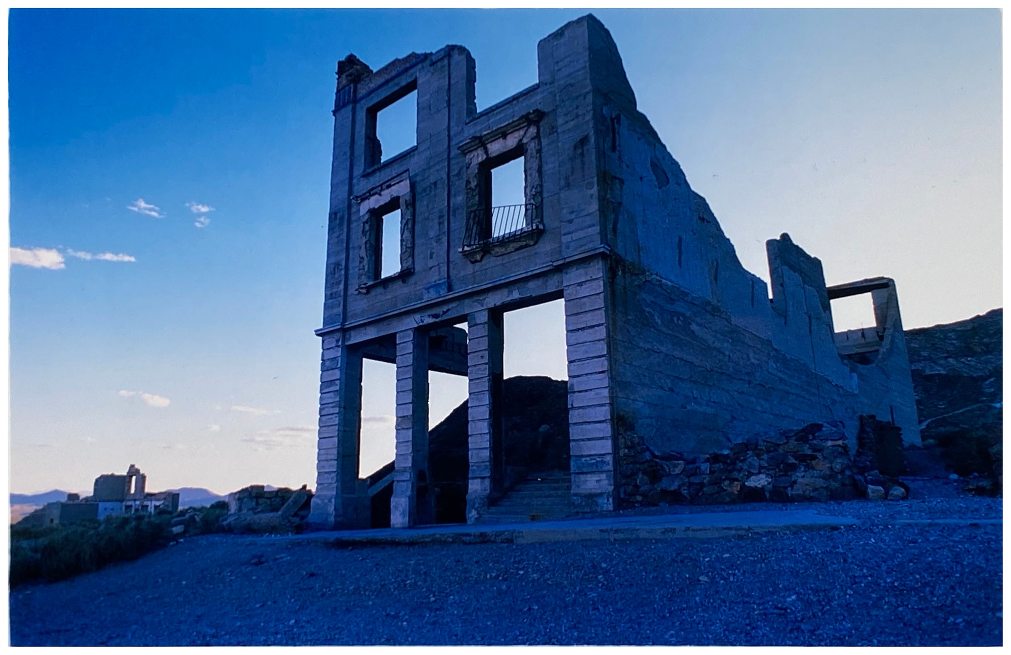 Photograph by Richard Heeps. The remnants of a rectangular building sits alone, surrounded by rubble and gravel in a blue light.