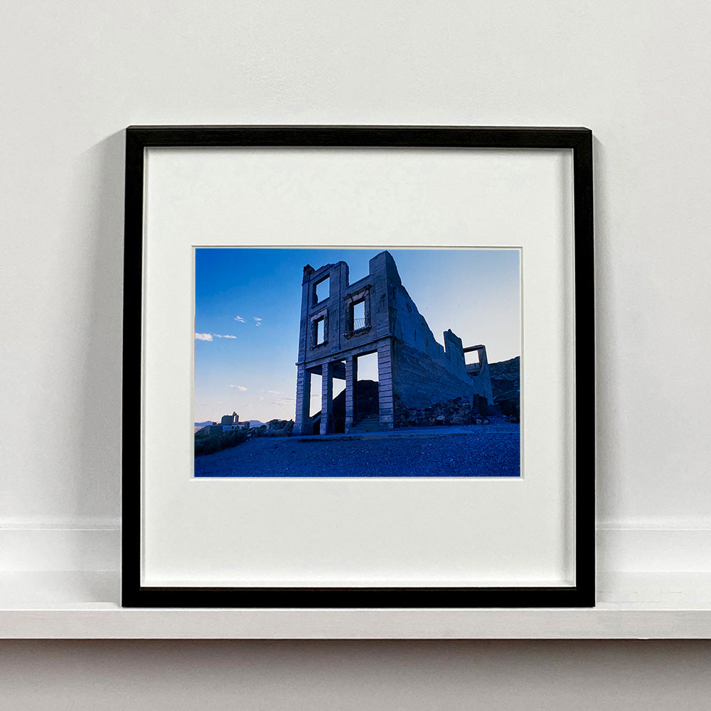 Black framed photograph by Richard Heeps. The remnants of a rectangular building sits alone, surrounded by rubble and gravel in a blue light.