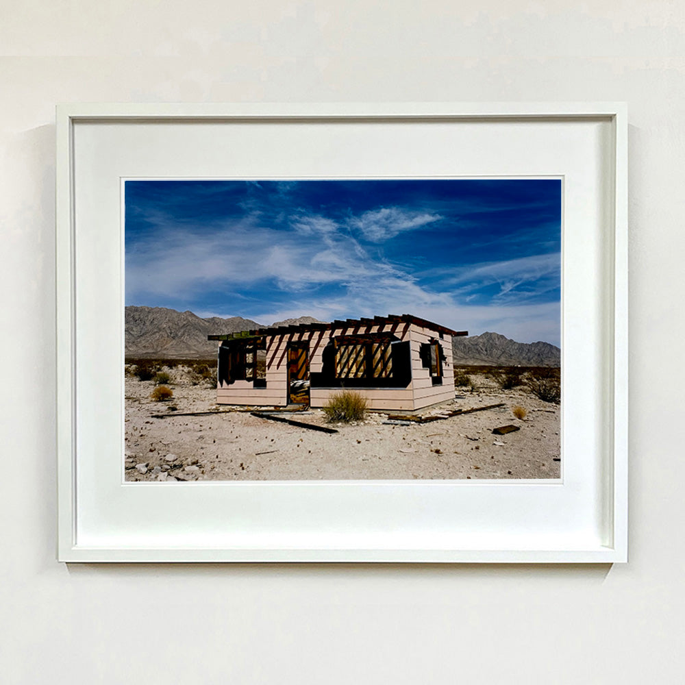 White framed photograph by Richard Heeps. An abandoned building sitting alone in a desert, tumble weed on the ground and hills in the background. A blue sky with white clouds.