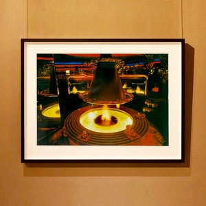 Black framed photograph by Richard Heeps. A fire pit burning inside a lounge bar. There is a conical hood over the fire and the fire is reflected in the metal and mirrors in the lounge.