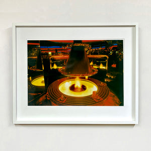 White framed photograph by Richard Heeps. A fire pit burning inside a lounge bar. There is a conical hood over the fire and the fire is reflected in the metal and mirrors in the lounge.