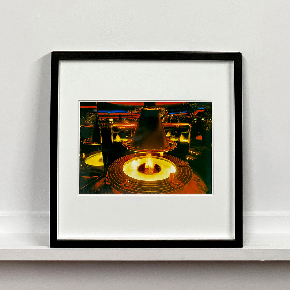 Black framed photograph by Richard Heeps. A fire pit burning inside a lounge bar. There is a conical hood over the fire and the fire is reflected in the metal and mirrors in the lounge.