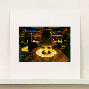 Mounted photograph by Richard Heeps. A fire pit burning inside a lounge bar. There is a conical hood over the fire and the fire is reflected in the metal and mirrors in the lounge.