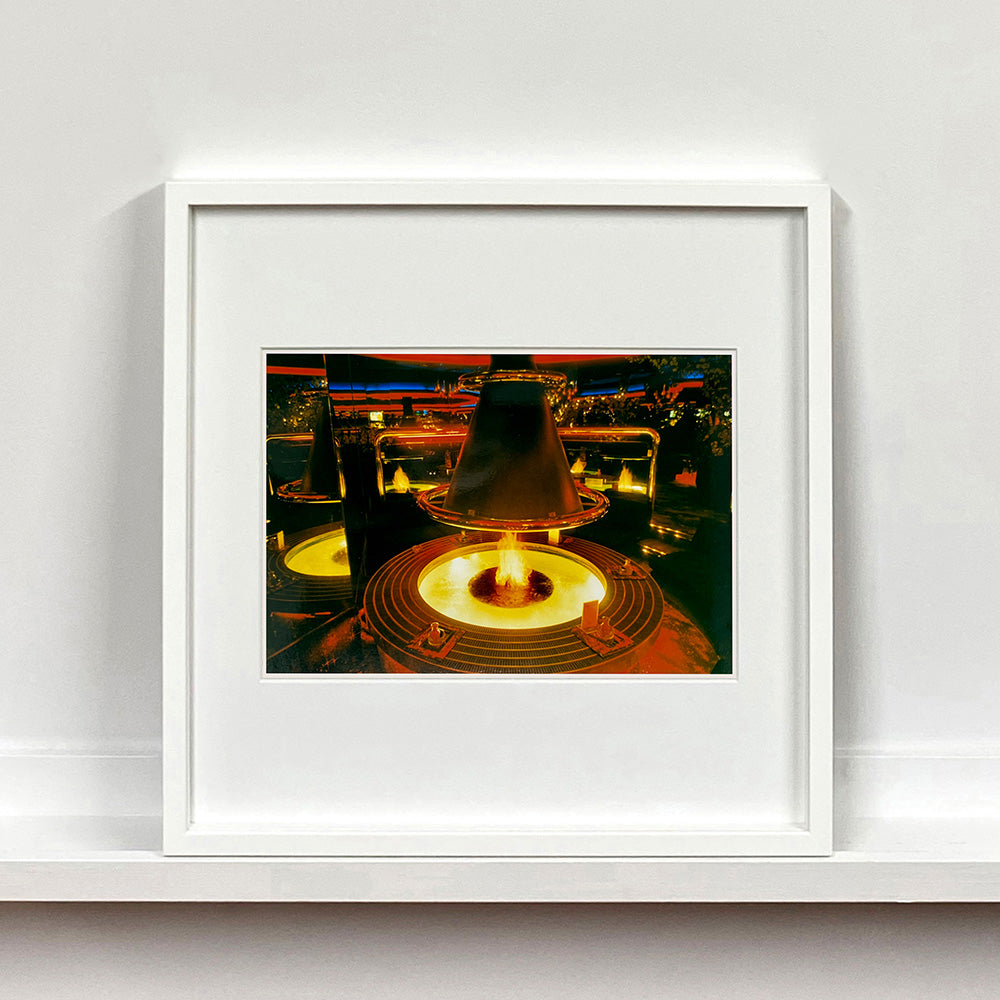 White framed photograph by Richard Heeps. A fire pit burning inside a lounge bar. There is a conical hood over the fire and the fire is reflected in the metal and mirrors in the lounge.