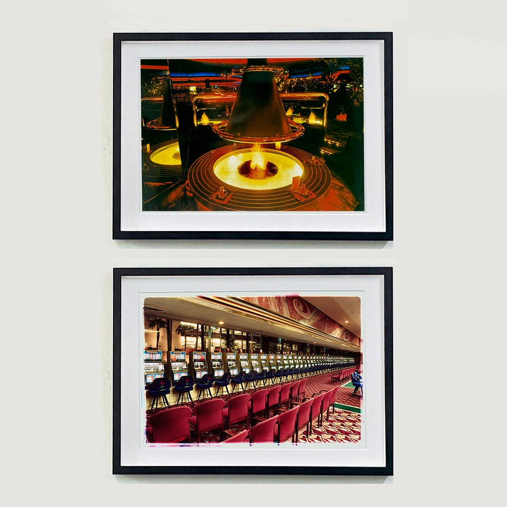 Two black framed photographs by Richard Heeps. The top photograph is of a fire pit burning inside a lounge bar. There is a conical hood over the fire and the fire is reflected in the metal and mirrors in the lounge. The bottom photograph is of a row of slot machines with two rows of red chairs lined up in front of them.
