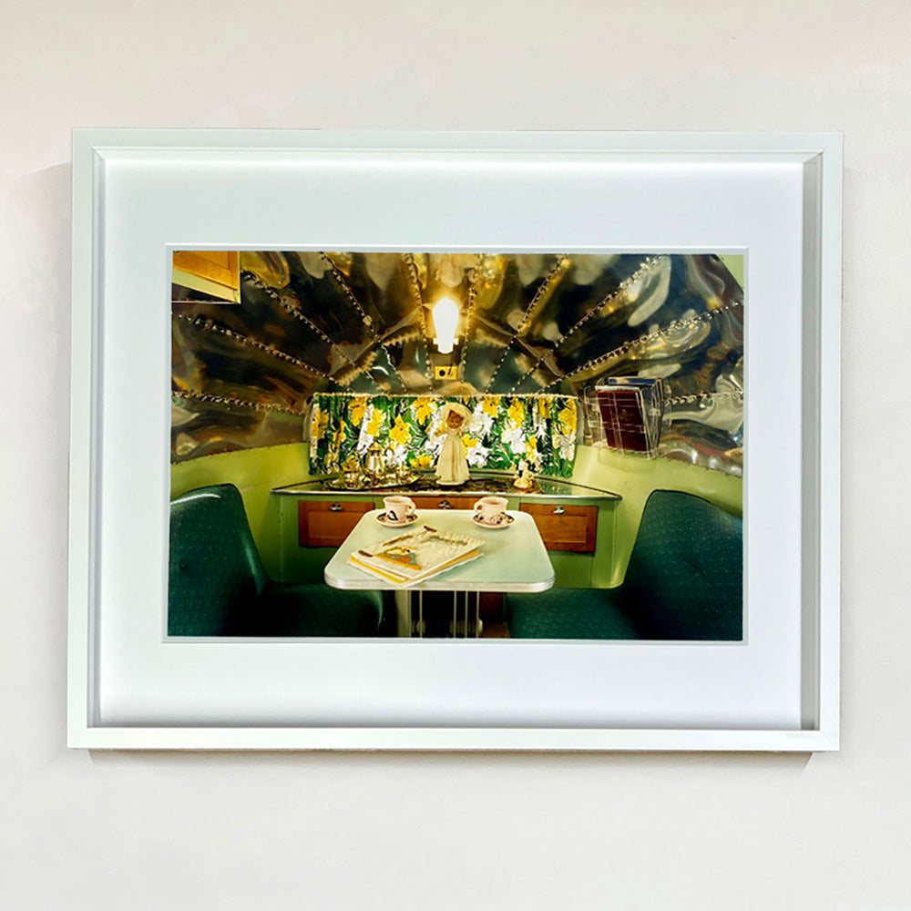 White framed photograph by Richard Heeps. Inside a trailer, there is a fixed table and chairs with two tea cups and saucers on the table. Behind on the shelf is a tea set and a doll dressed as a bride.
