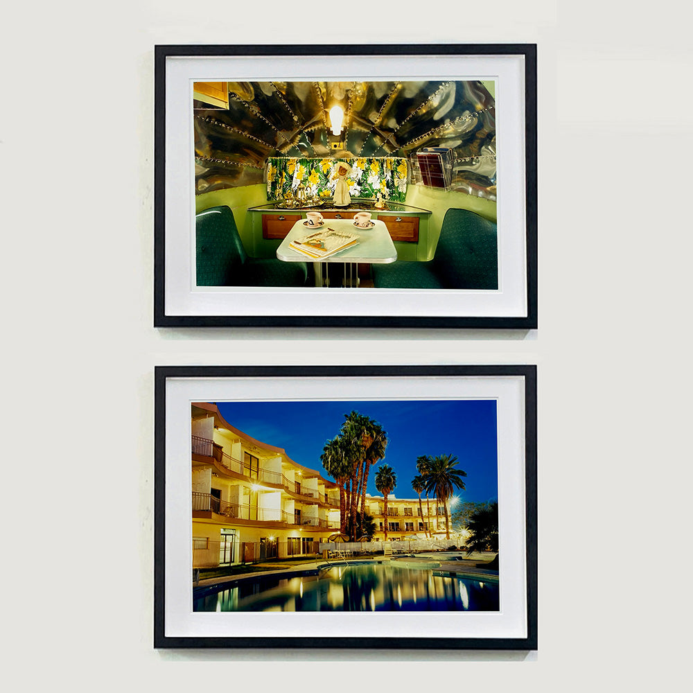 Two black framed photographs by Richard Heeps. The top photograph is inside a trailer, there is a fixed table and chairs with two tea cups and saucers on the table. Behind on the shelf is a tea set and a doll dressed as a bride. The bottom photograph is a hotel and its reflection in the swimming pool taken at night time, lit by the hotel lights.