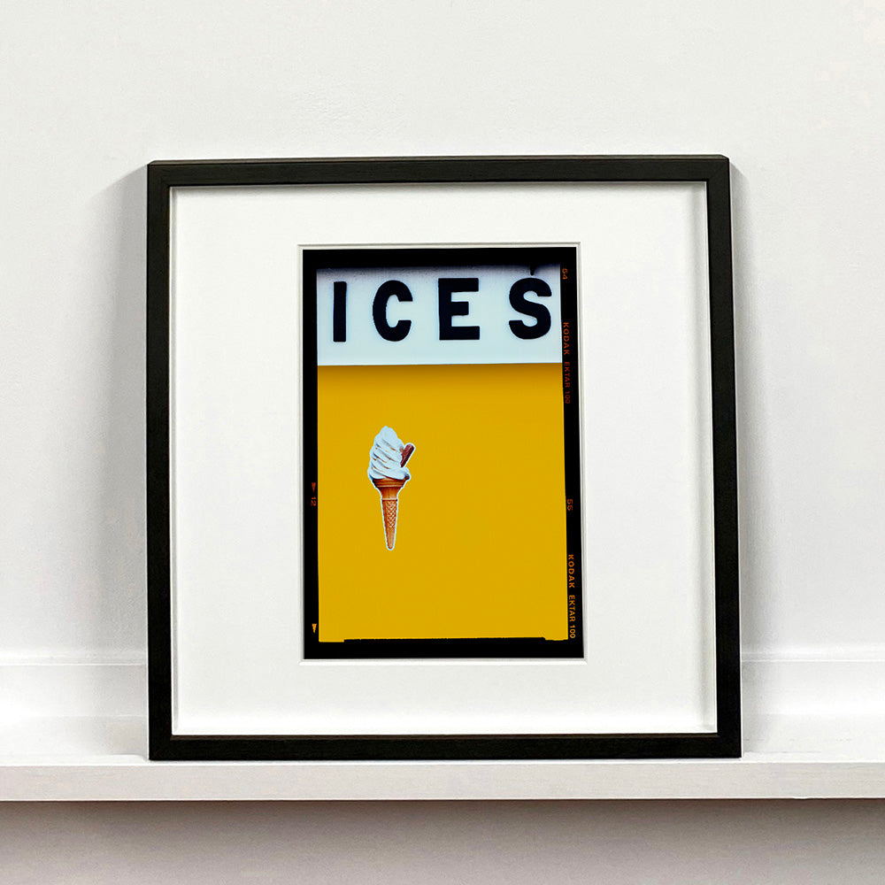 Black framed photograph by Richard Heeps.  At the top black letters spell out ICES and below is depicted a 99 icecream cone sitting left of centre against a mustard yellow coloured background.  