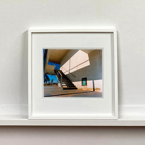 White framed photograph by Richard Heeps. The photograph has a metal staircase on the outside of a cream colour motel. The staircase has a ceiling but no sides so leads to blue sky. Behind the building is a blue sky and palm trees of the Californian Desert.