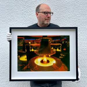 Black framed photograph held by photographer Richard Heeps. A fire pit burning inside a lounge bar. There is a conical hood over the fire and the fire is reflected in the metal and mirrors in the lounge.