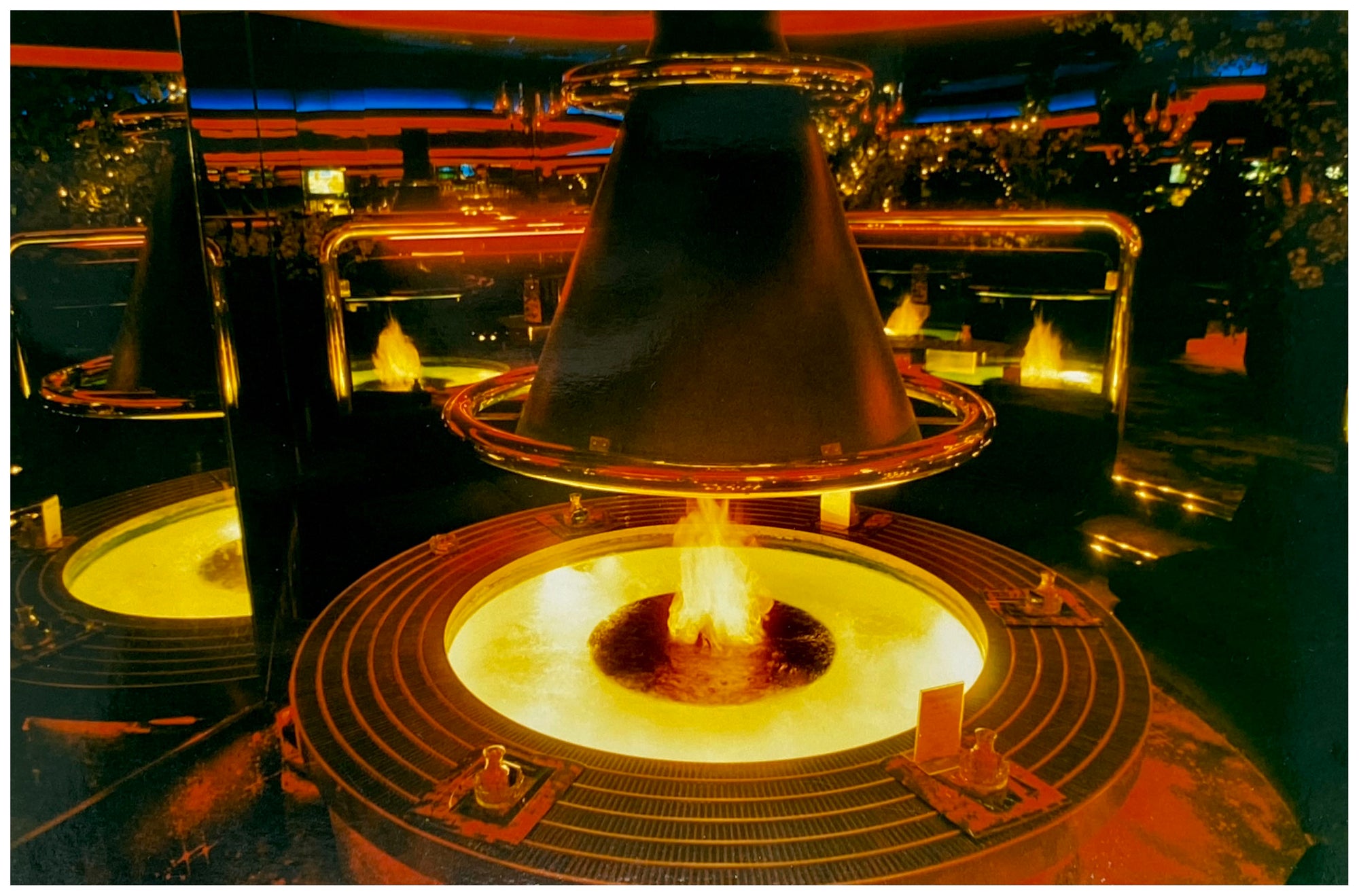Photograph by Richard Heeps. A fire pit burning inside a lounge bar. There is a conical hood over the fire and the fire is reflected in the metal and mirrors in the lounge.