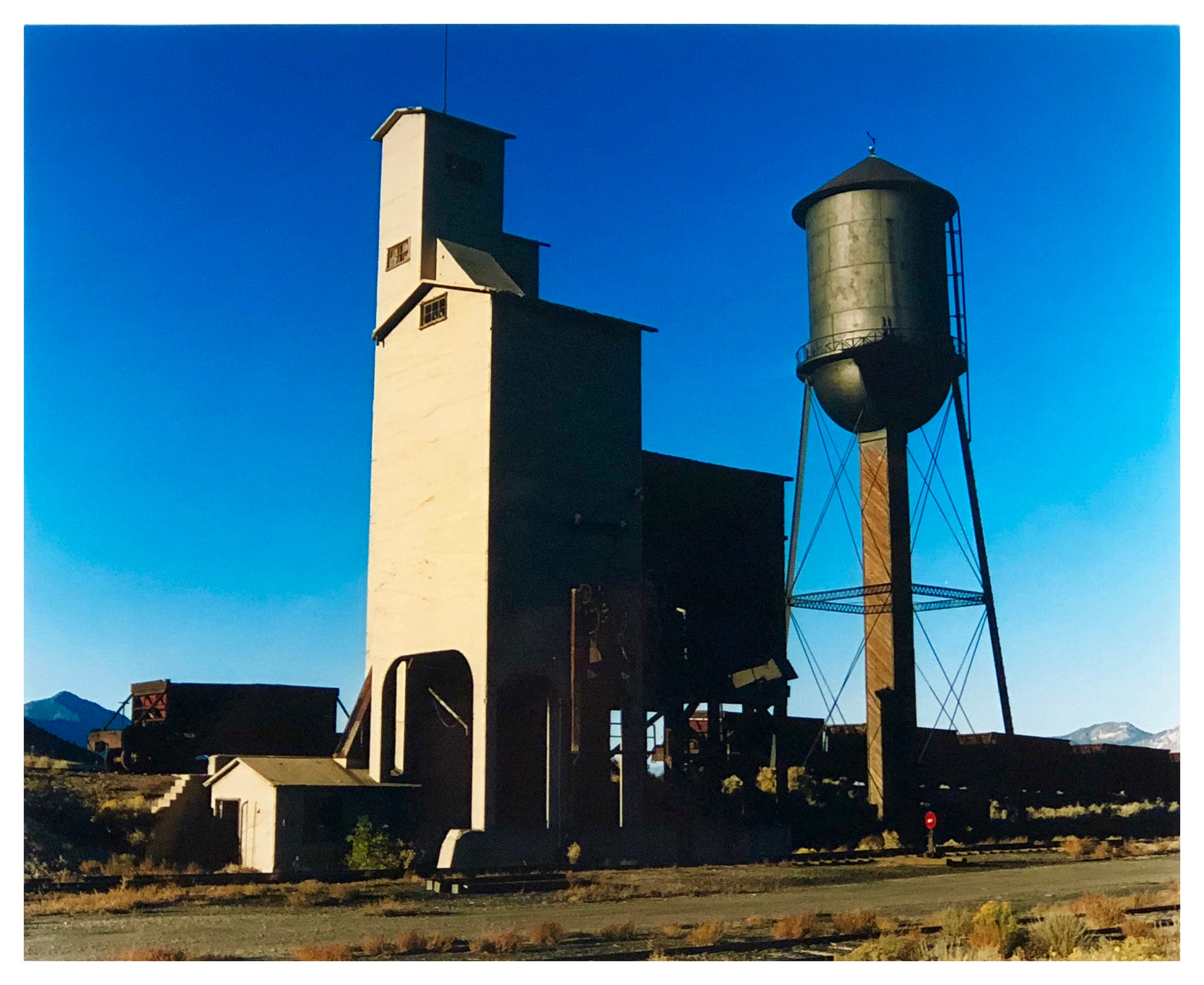 A grain silo and a water tower against a blue sky in Ely, Nevada, iconic American architecture photography by Richard Heeps.
