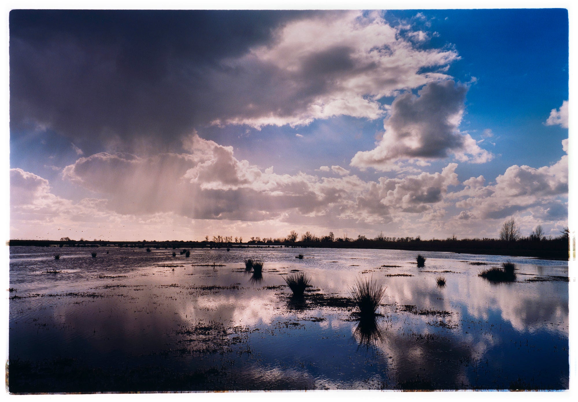Photograph by Richard Heeps. Fenland expanse with water and tufts of grass and an expansive fenland sky, blue with white and grey clouds reflected in the water below.