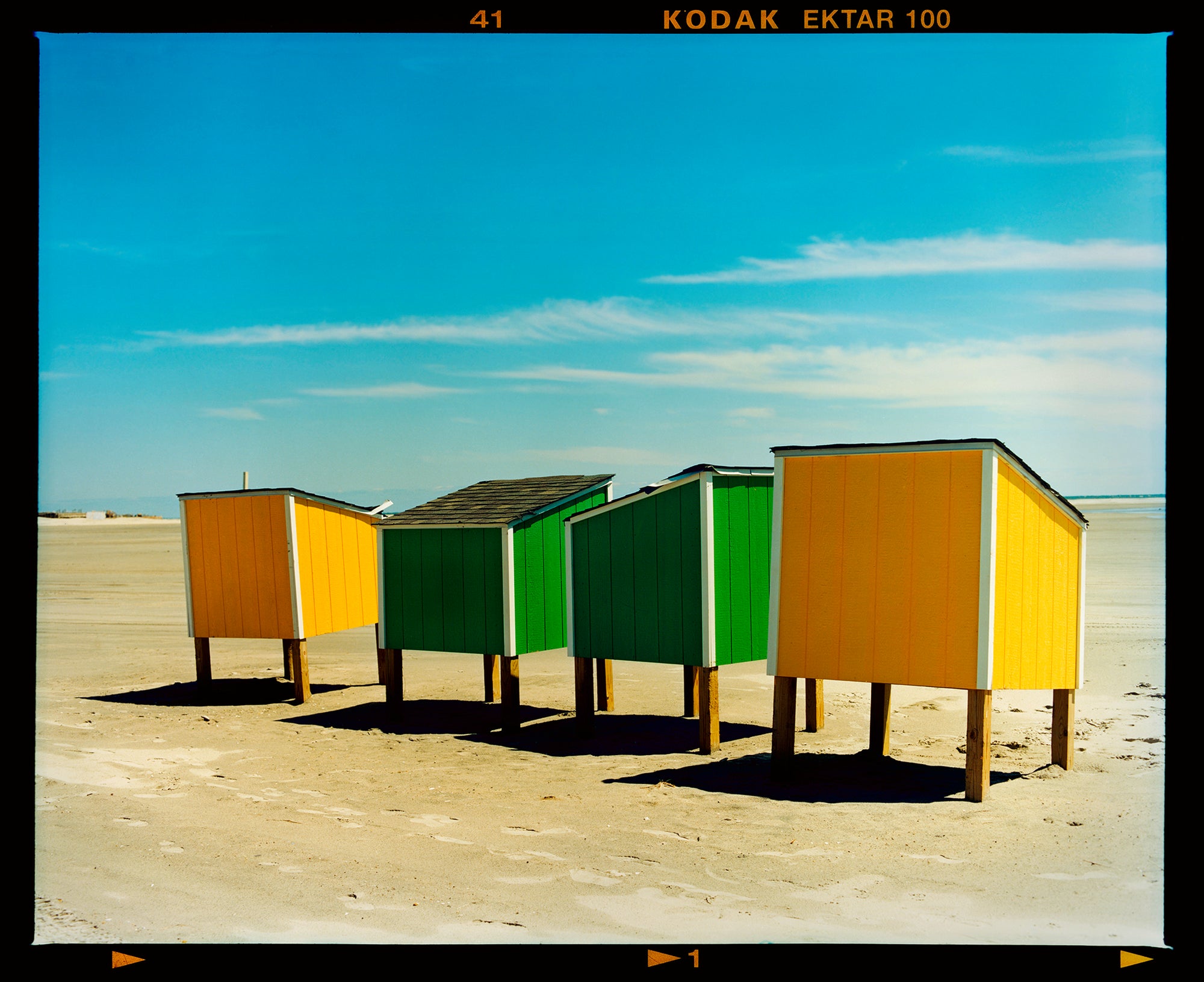 Photograph by Richard Heeps. Yellow and green beach lockers stand on their wooden legs on the clear sandy beach.
