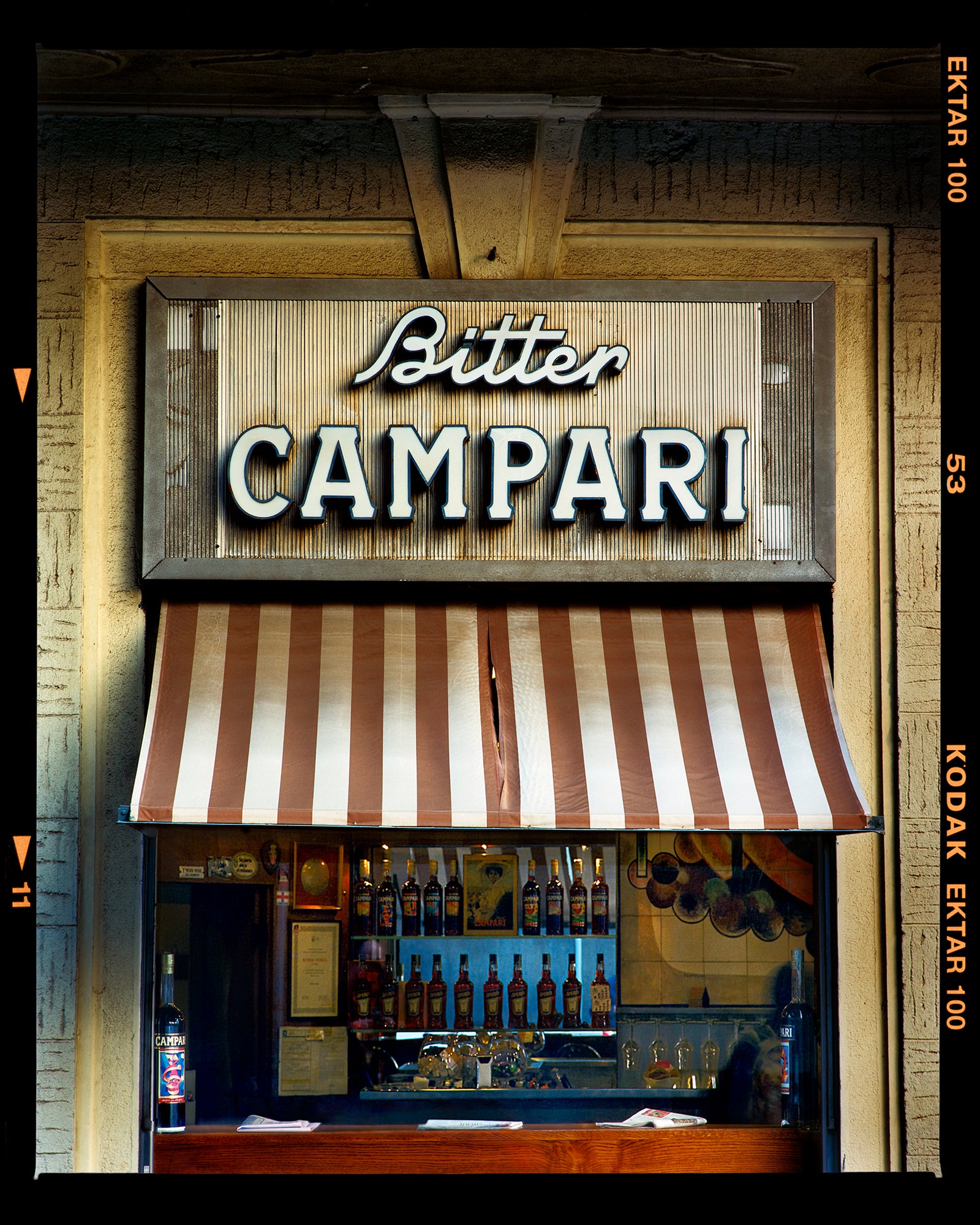 Photograph by Richard Heeps. A shop selling drinks, it has a red and white awning and the shop is called Bitter Campari in white lettering. This photograph has a black rebate.