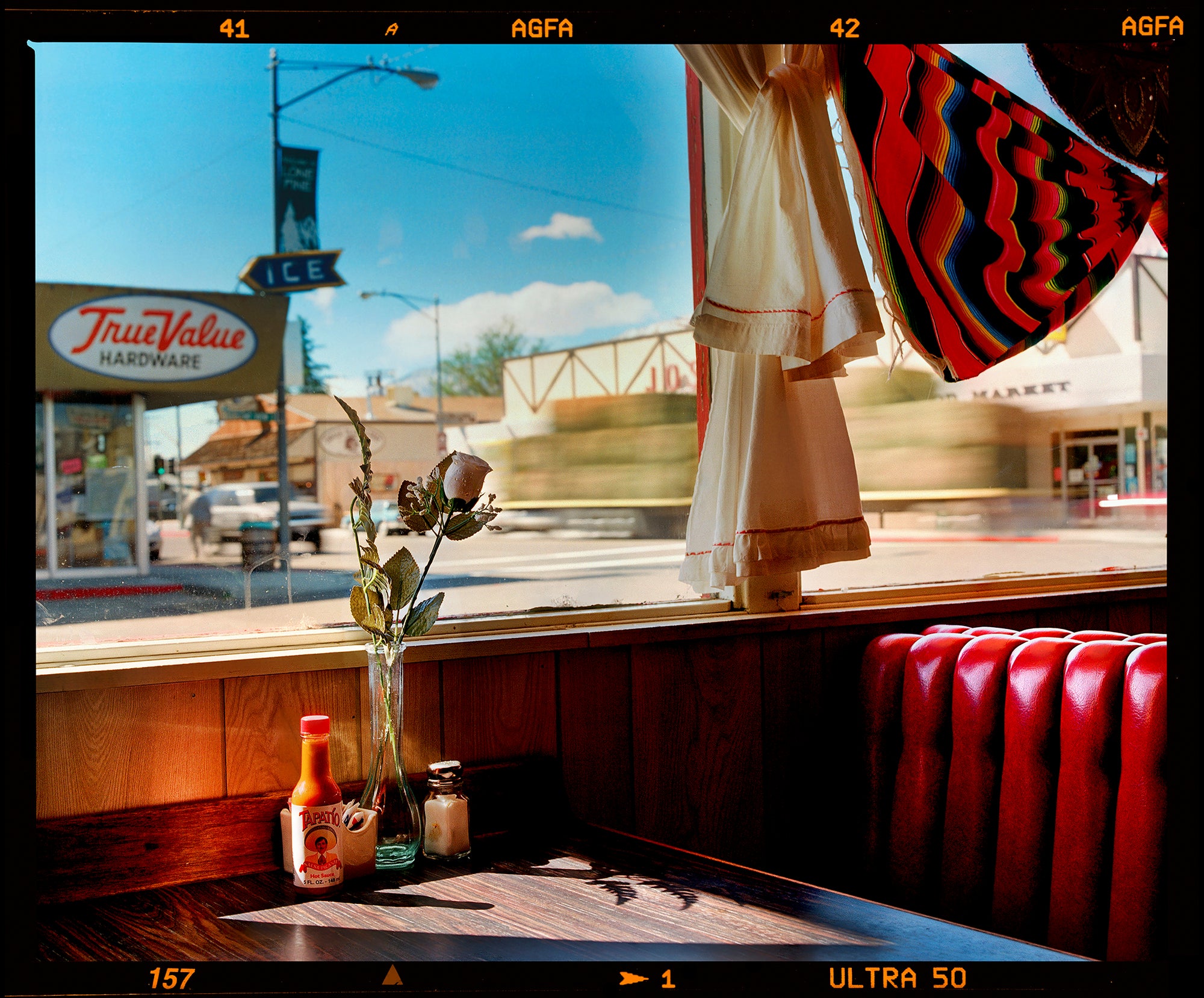 Photograph by Richard Heeps. Inside an American diner the light is shining on the table and the seat is the classic vibrant red faux-leather of a retro diner. The photograph is looking out of the window into the outside street.