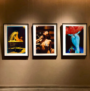 Three framed art prints featuring colorful artistic burlesque photographs by Richard Heeps, displayed side-by-side on a dark brown gallery wall under spotlights.