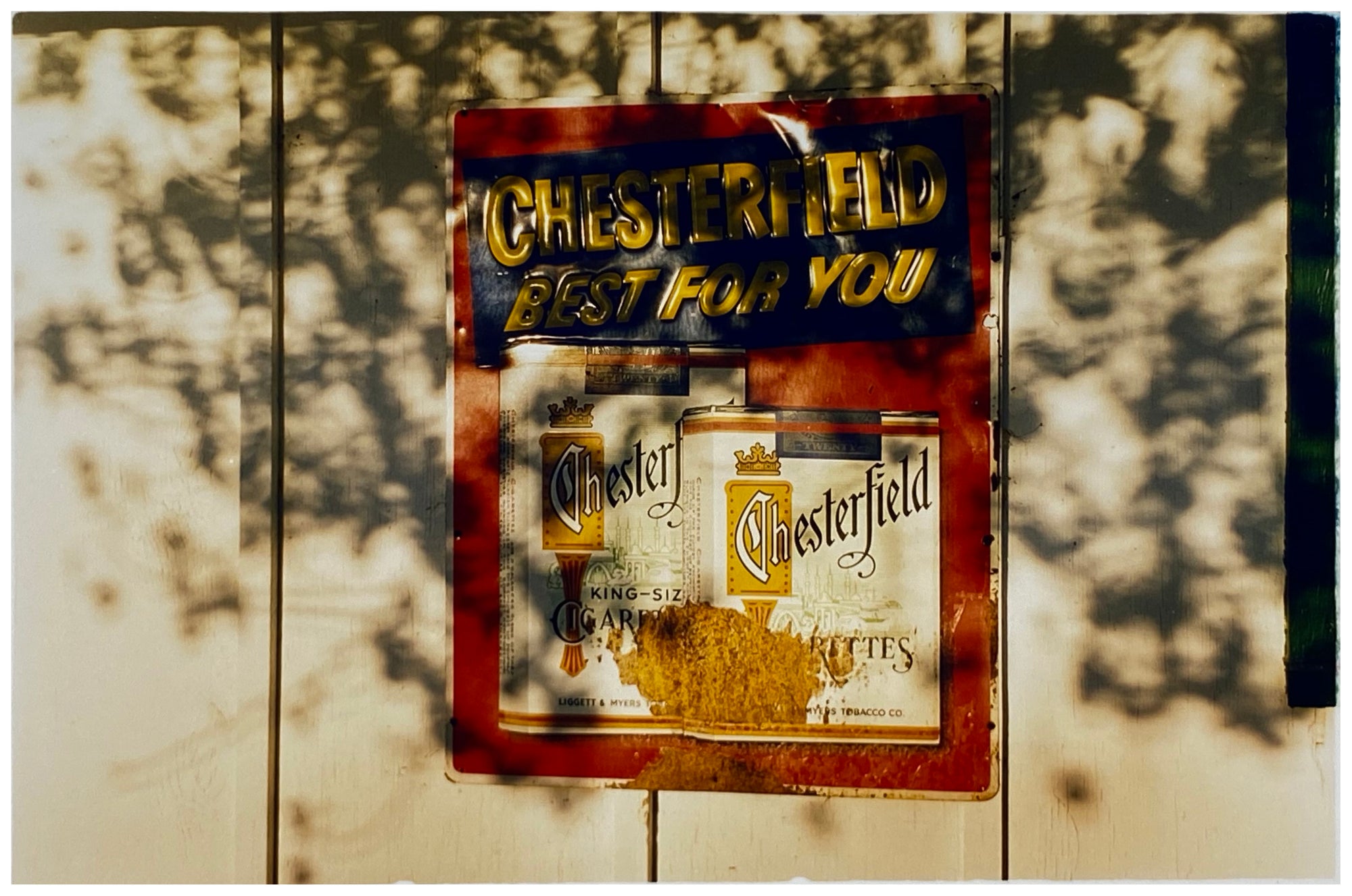 Photograph by Richard Heeps. A plastic retro cigarette sign  for Chesterfield cigarettes is stuck to a white wash wooden fence. The words Chesterfield Best for You are raised on the sign and the sign is in a dappled light.