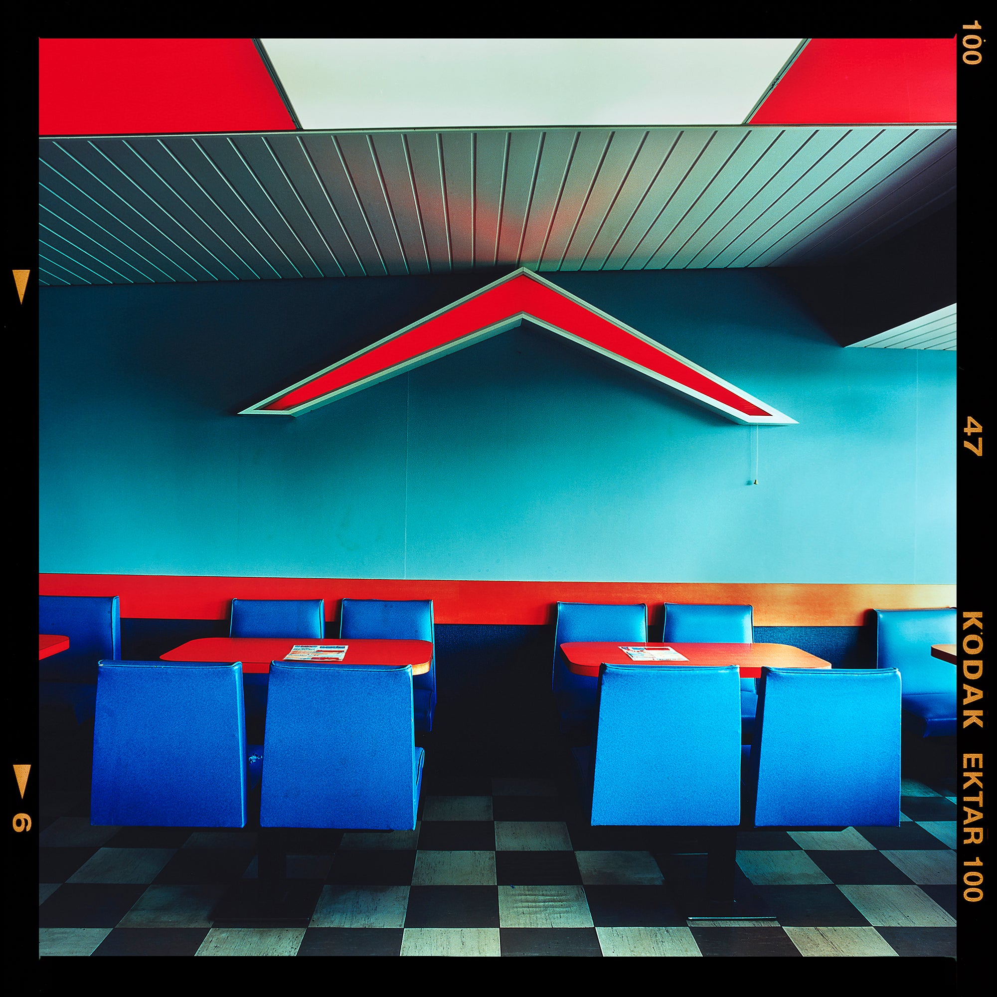 Photograph by Richard Heeps. This photograph captures the inside of a Wimpy Restaurant in Norfolk. There is bright blue seats and red tables. The walls are blue and there is a big red chevron light attached to the wall. 
