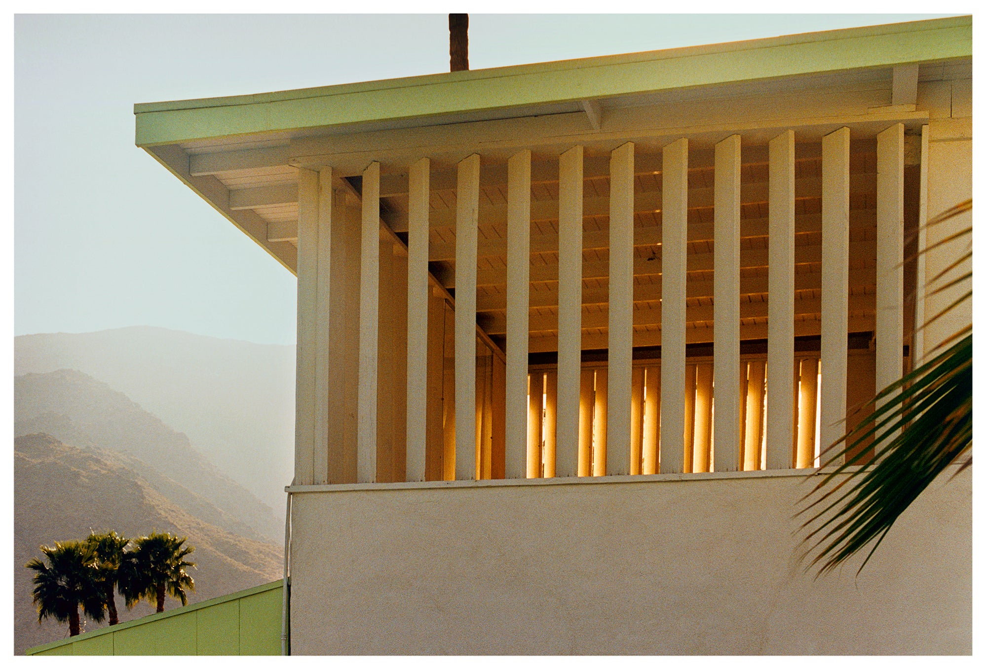 Photograph by Richard Heeps.  The corner of the top of a cream colour building with big windows and vertical slats, on the left hand side are mountains bathing in the mist of dawn.