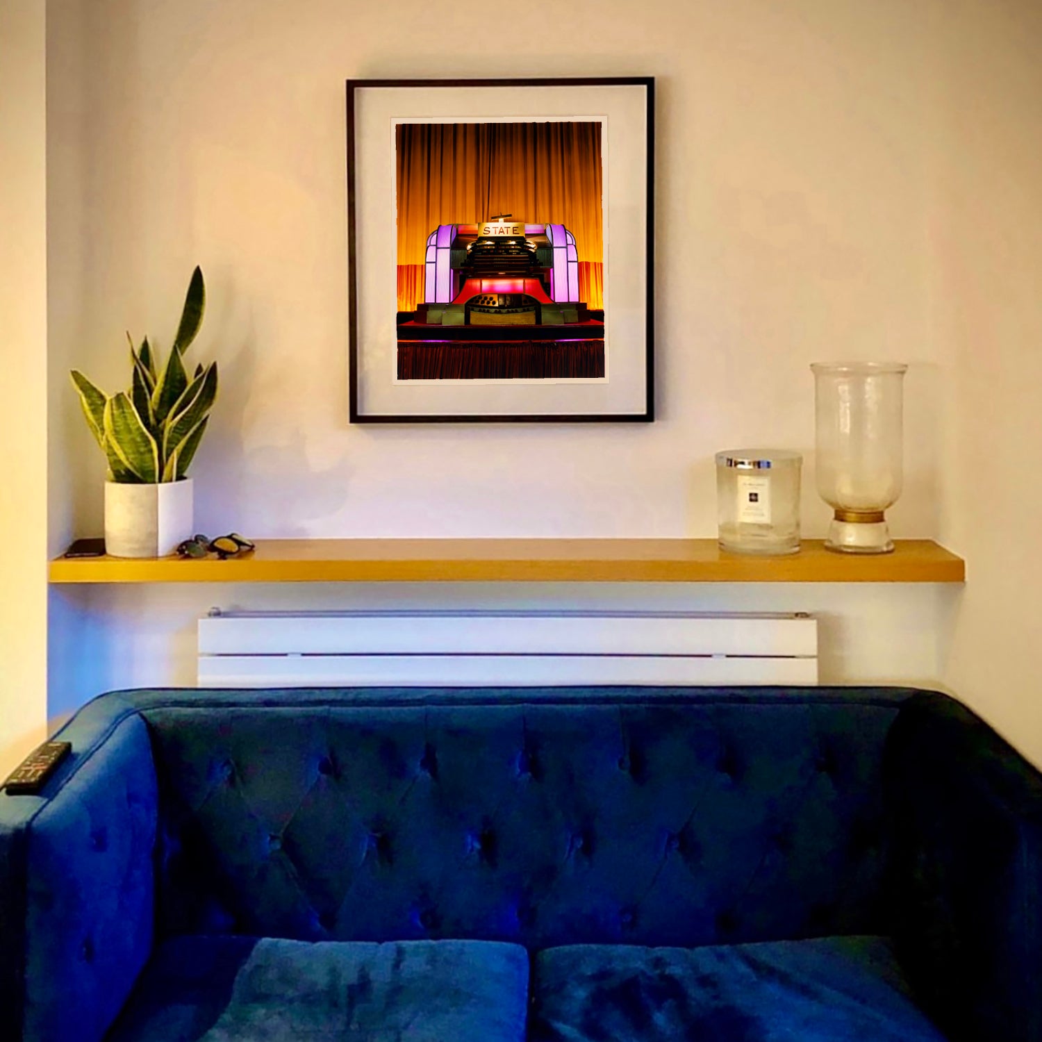 Framed artwork Compton Organ The State above a blue sofa with a plant and candle on a shelf.