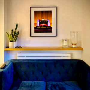 Framed artwork Compton Organ The State above a blue sofa with a plant and candle on a shelf.
