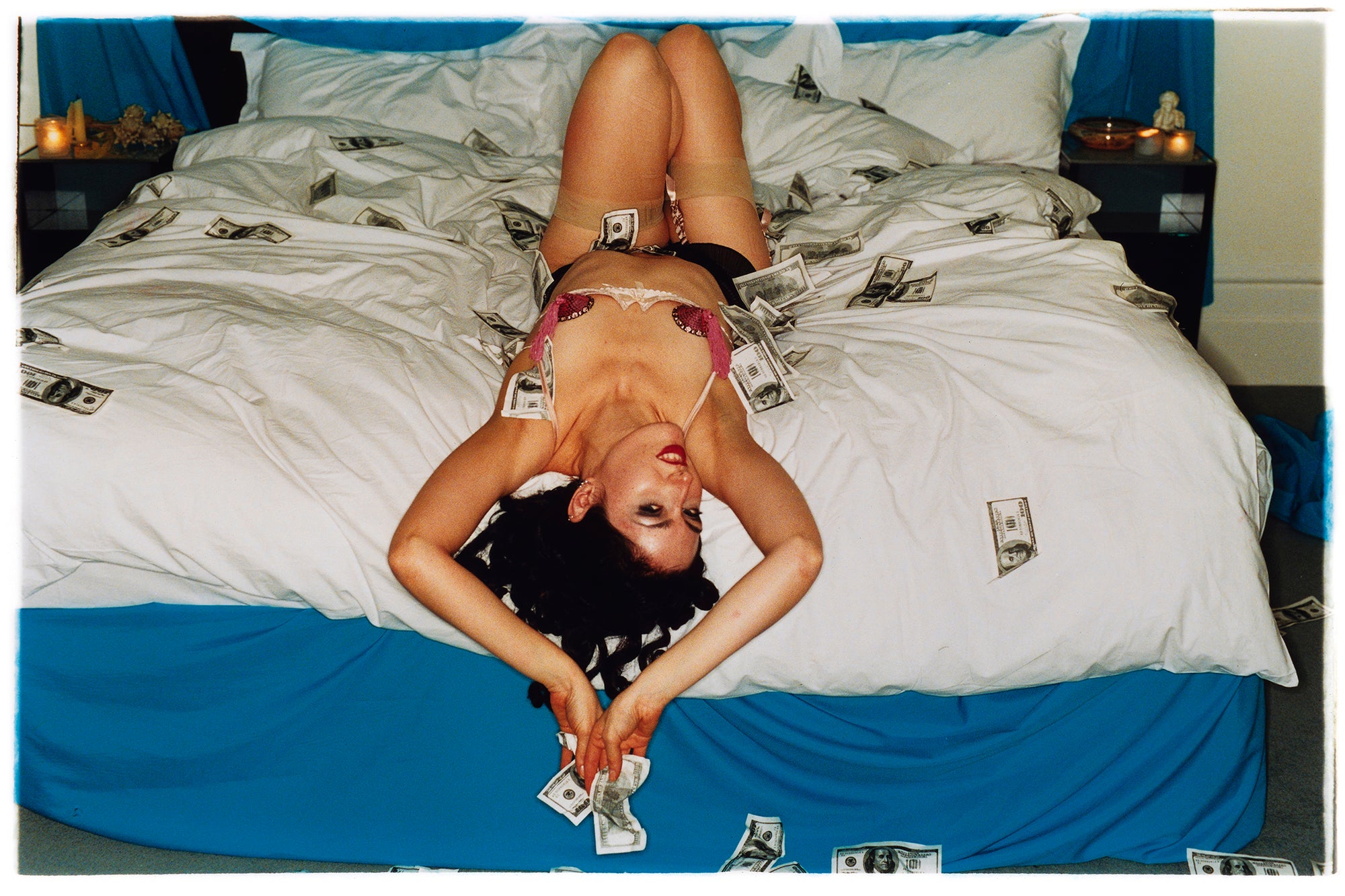 Photograph by Richard Heeps. A woman lies semi naked on a bed with money surrounding her. The photograph is taken from the end of the bed so the woman appears upside down. The bed has a white duvet with blue under sheets.