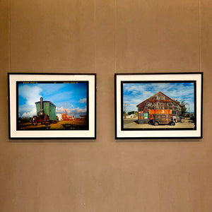Two framed landscape photographs by Richard Heeps hanging side-by-side on a beige wall. The left frame shows a green steam train on a track under a blue sky, and the right frame shows an old weathered barn with a rusted orange truck under a blue sky.