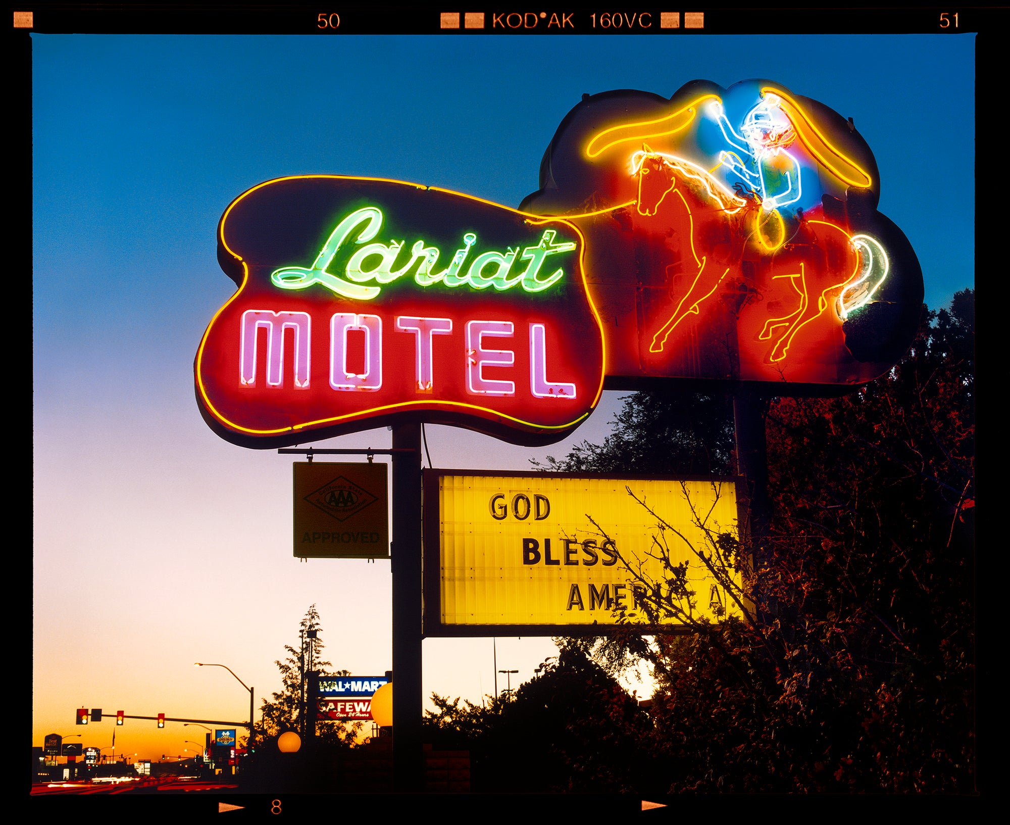 Photograph by Richard Heeps. The photograph is taken in the evening, a neon sign belonging to the Lariat Motel, with a cowboy on horseback with a lasso on the right of the words, half way down the sign post is the yellow sign God Bless America.