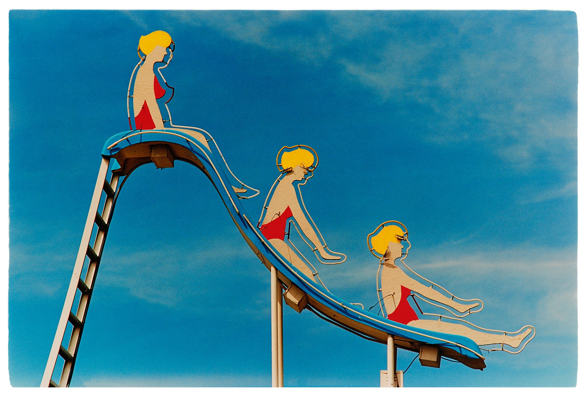 Photograph by Richard Heeps. A neon sign against a blue sky in Las Vegas featuring three women with red swimming costumes and blond hair on a slide. 