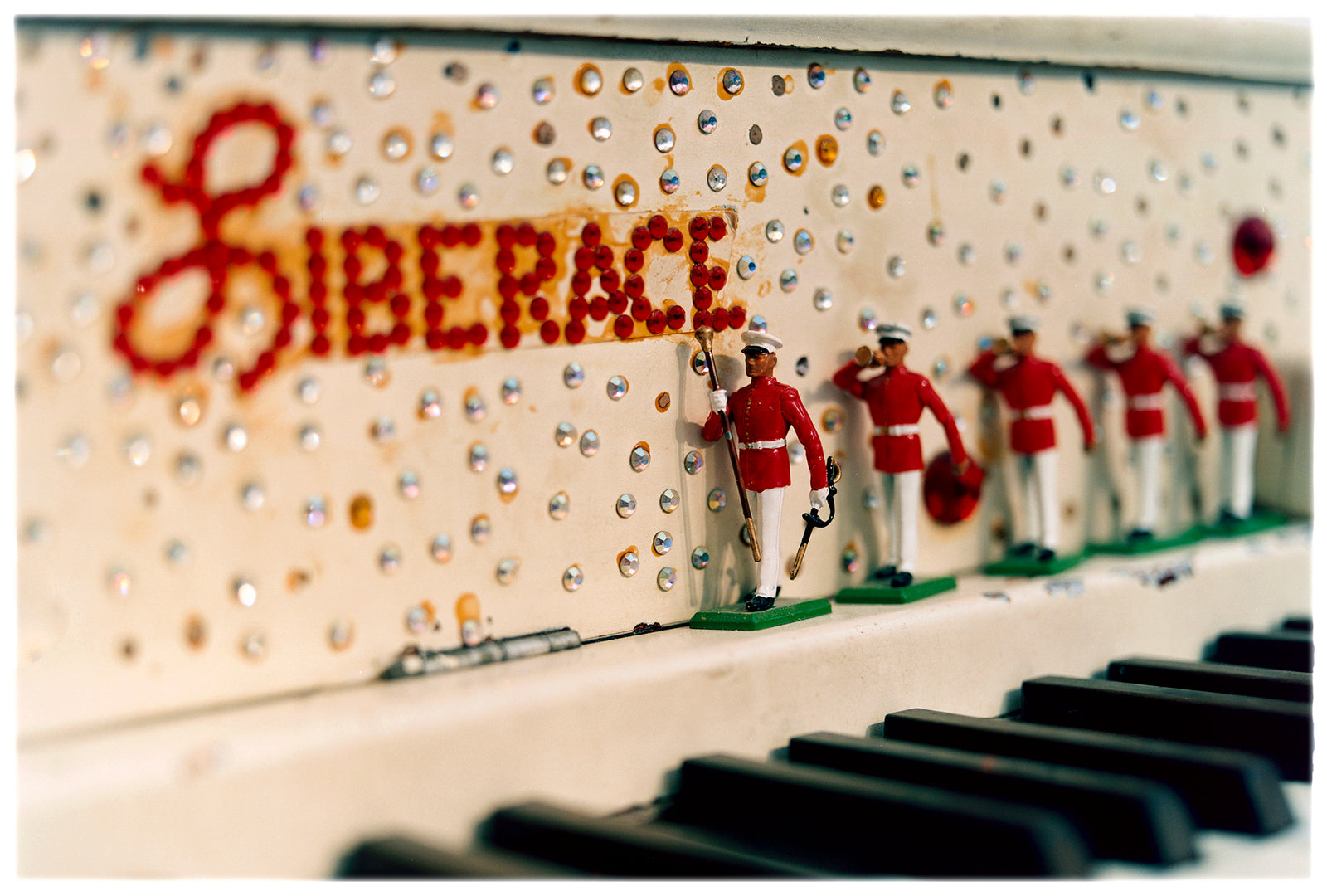 Photograph by Richard Heeps. A close up of Liberace's piano. This captures the black keys and the fall board which is decorated with crystals and red buttons spelling out Liberace in capitals. There are also 5 red and white metal soldiers positioned marching along the rim.