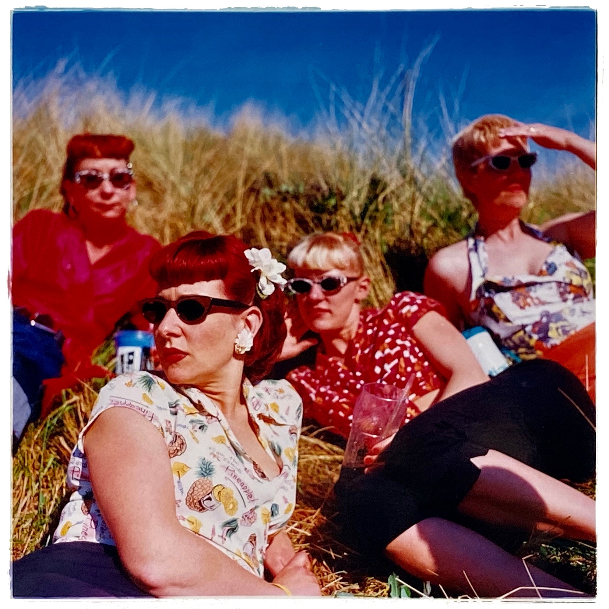 Photograph by Richard Heeps. A photo of four women dressed in a retro style all wearing sunglasses on a sunny day lounging on a hill with high grass.