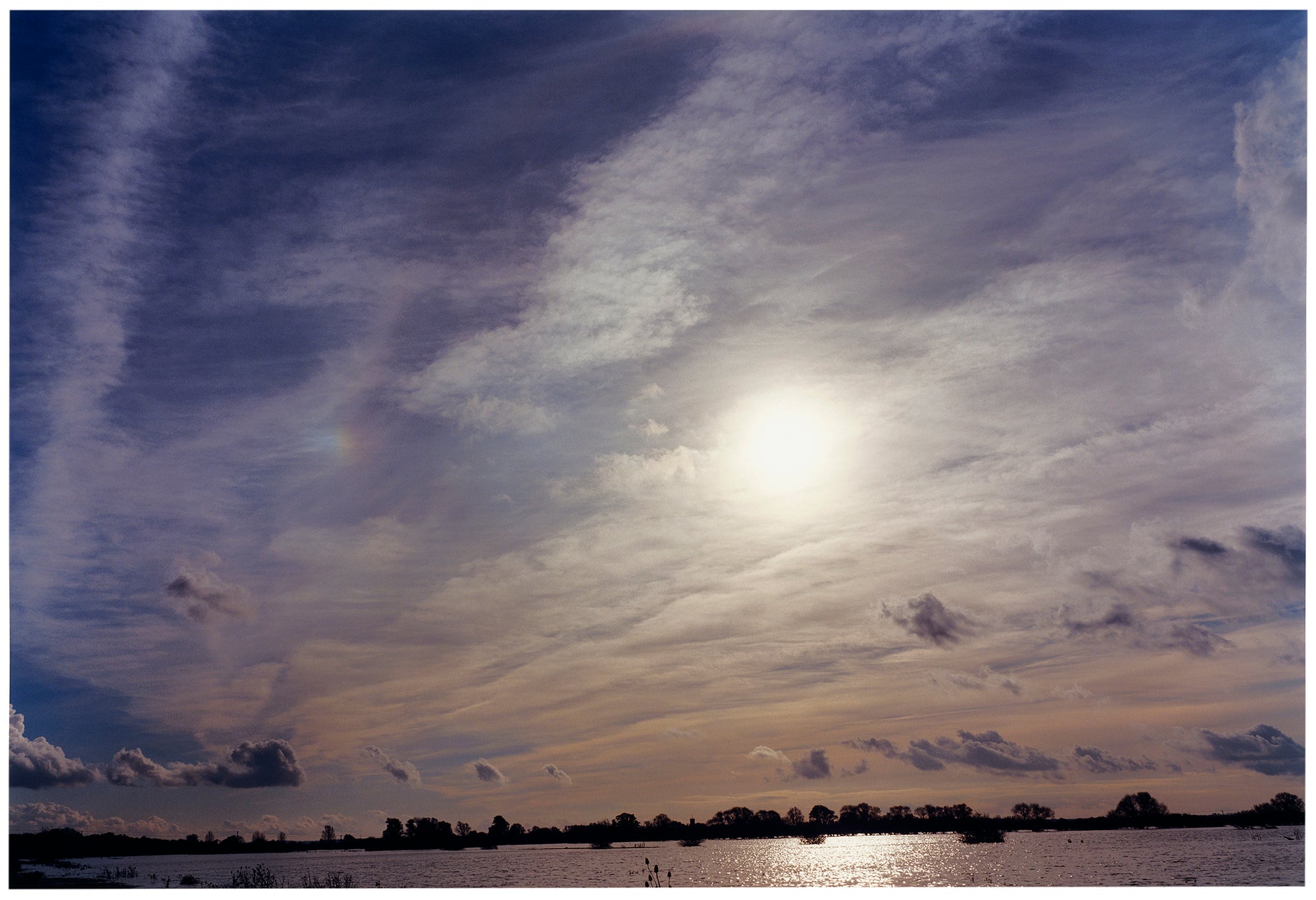 Photograph by Richard Heeps. A big sky with exciting clouds, an obscured sun and a flood of water features at the bottom reflecting the sun's light.