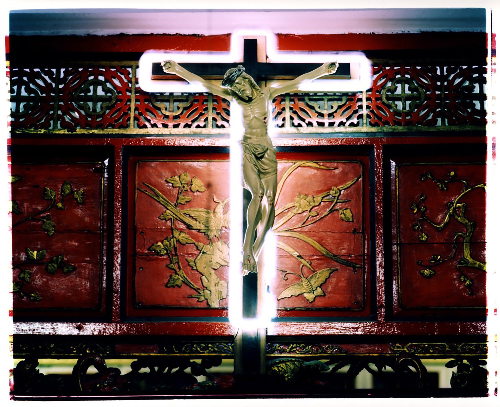 Photograph by Richard Heeps. A marble effect Jesus Christ statue hangs on a wooden cross which is surrounded with a white neon light. This sits against a wooden screen with gold carved patterns of flowers and butterlies.
