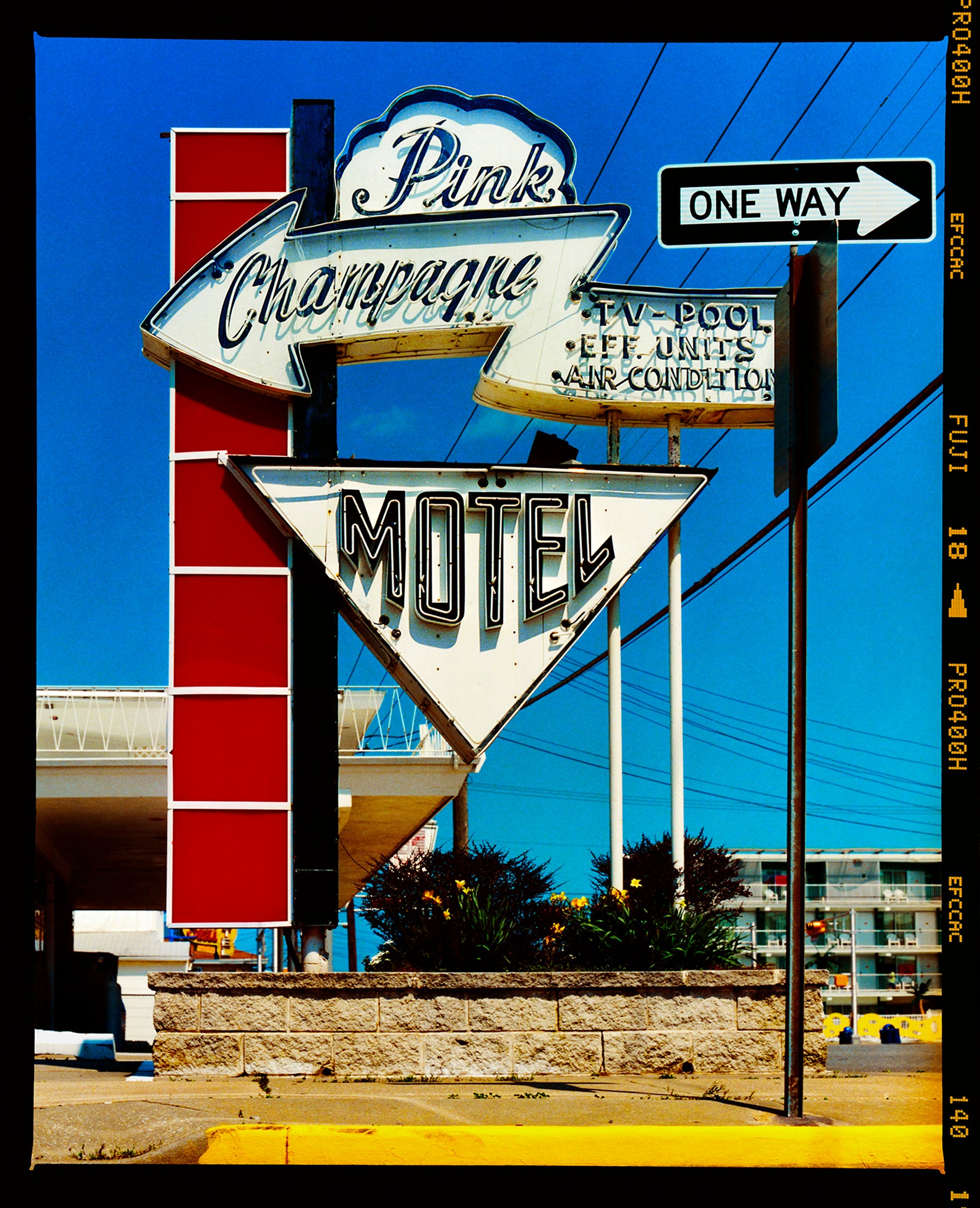 Photograph by Richard Heeps. A group of signs on an American Road sitting outside a motel mounted on a post with a vertical line of red squares.  The top of  the sign shows an arrow pointing to the left to Pink Champagne, there is an arrow pointing down with MOTEL written in it. In front of this motel sign is a one way sign pointing to the right. 
