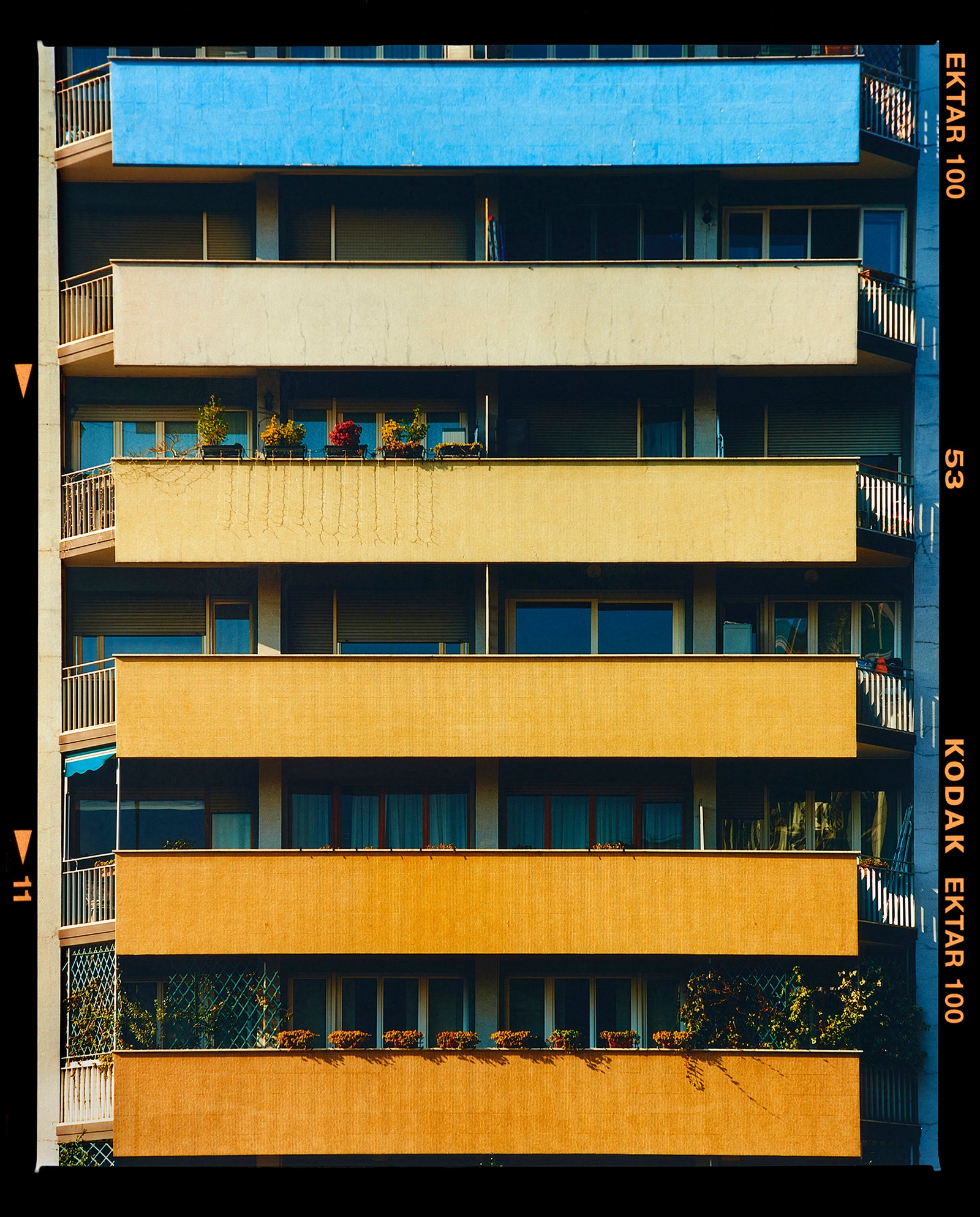 Photograph by Richard Heeps. Photograph of an apartment building with coloured balconies, blue at the top balcony and then fading from light yellow to a sunburnt yellow at the bottom.