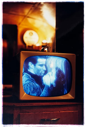 Photograph by Richard Heeps. A retro small brown television sits on a wooden table displaying a black and white film which shows as blue and white. In the background of the television is a warm light.