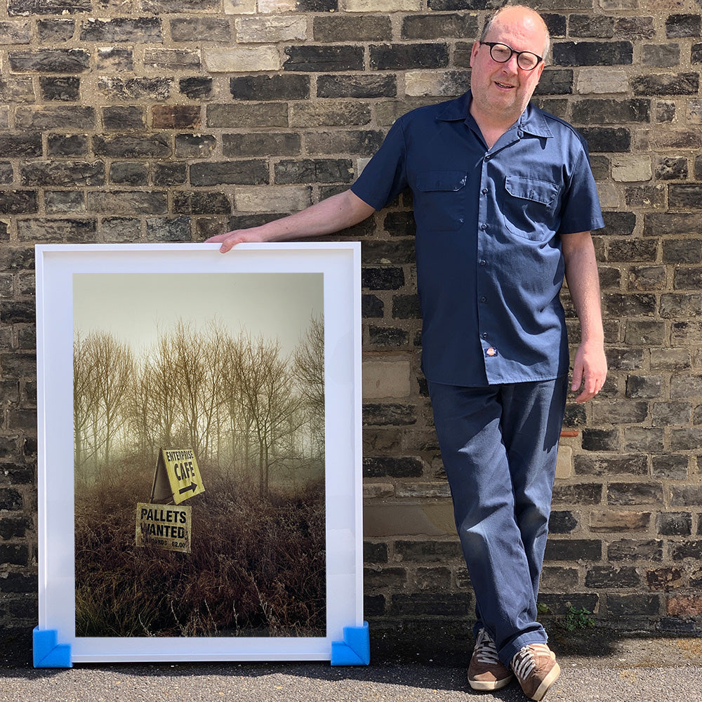 White framed photograph held by photographer Richard Heeps.  A roadside sign for The Enterprise Cafe and Pallets Wanted sit in brown shrub with wintry trees and sky in the background.