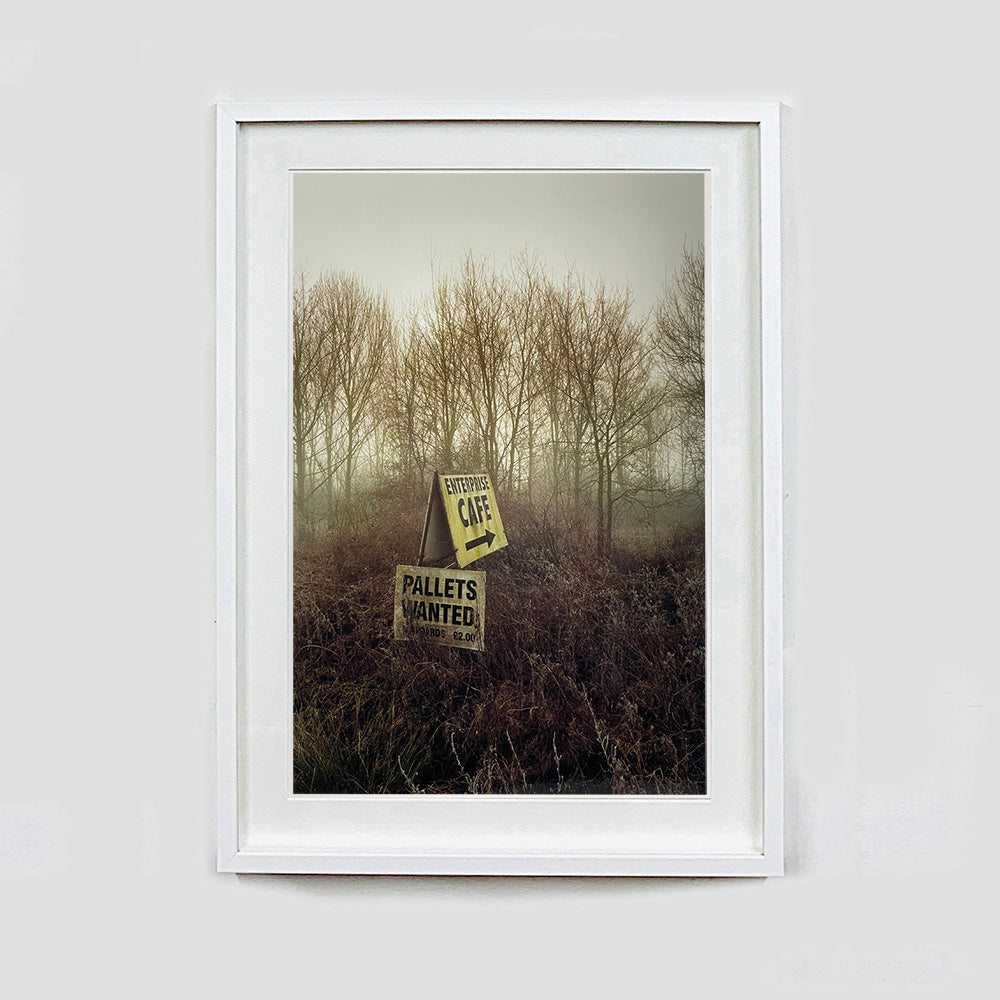 White framed photograph by Richard Heeps.  A roadside sign for The Enterprise Cafe and Pallets Wanted sit in brown shrub with wintry trees and sky in the background.