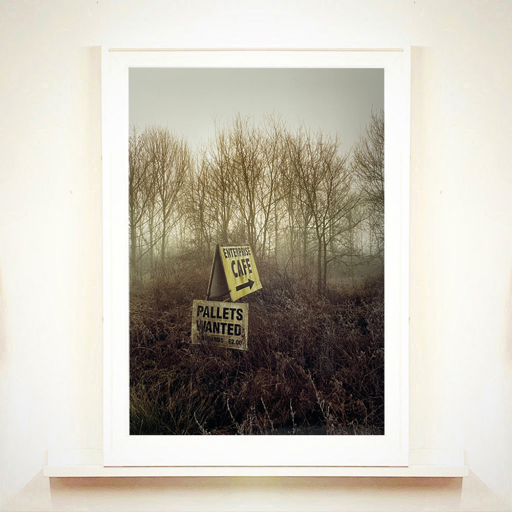 White framed photograph by Richard Heeps.  A roadside sign for The Enterprise Cafe and Pallets Wanted sit in brown shrub with wintry trees and sky in the background.