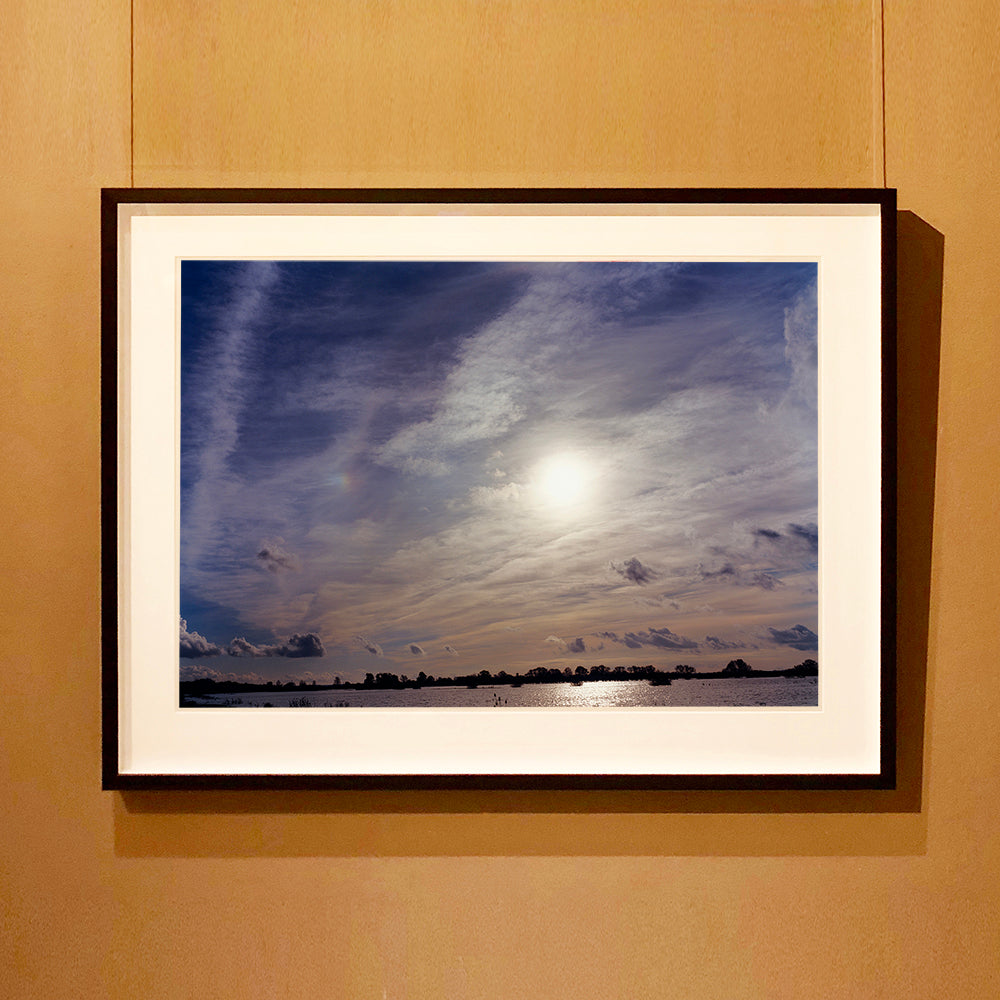Black framed photograph by Richard Heeps. A big sky with exciting clouds, an obscured sun and a flood of water features at the bottom reflecting the sun's light.