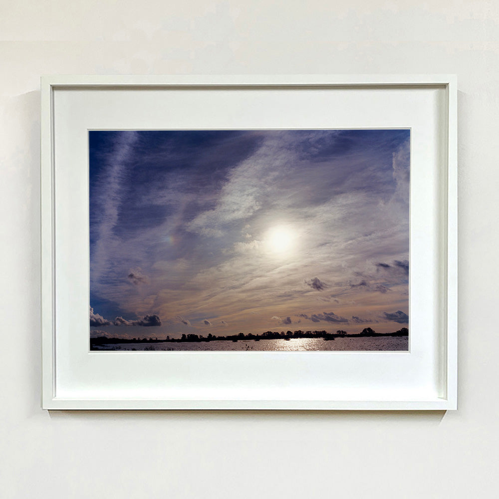 White framed photograph by Richard Heeps. A big sky with exciting clouds, an obscured sun and a flood of water features at the bottom reflecting the sun's light.