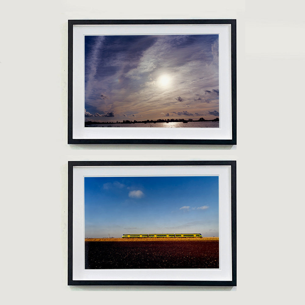 Two black framed photographs by Richard Heeps. At the top is a photograph of a  big sky with exciting clouds, an obscured sun and a flood of water features at the bottom reflecting the sun's light. The photograph at the bottom features a yellow train moving through the fen countryside from left to right.