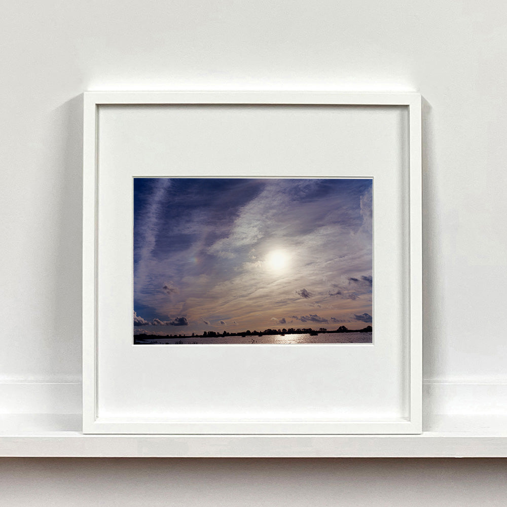 White framed photograph by Richard Heeps. A big sky with exciting clouds, an obscured sun and a flood of water features at the bottom reflecting the sun's light.