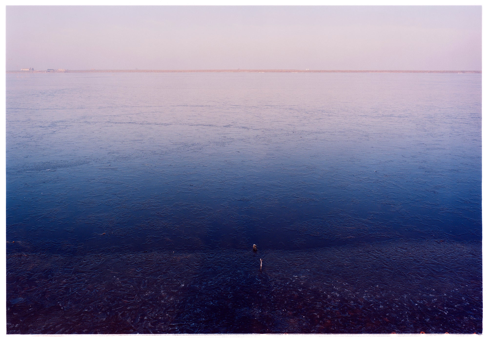 Photograph by Richard Heeps. This photograph is looking towards the water. From a distance you can see two blocks of colour, blue at the bottom and lilac at the top, closer up you see the move of the water and a thin land strip along the horizon.