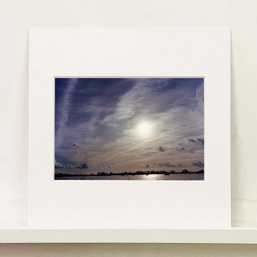 Mounted photograph by Richard Heeps. A big sky with exciting clouds, an obscured sun and a flood of water features at the bottom reflecting the sun's light.